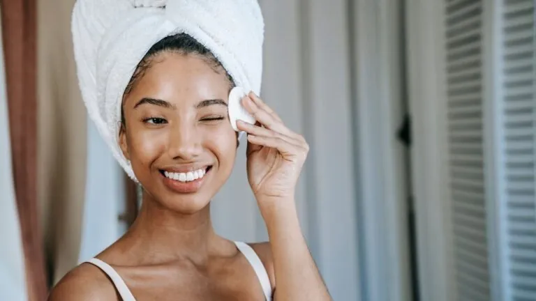 Smiling woman in a hair towel wiping her face with a cotton pad