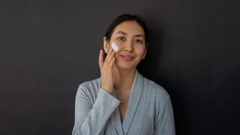 Woman in a grey robe applying moisturiser to her cheek